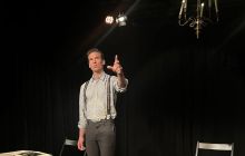 Actor Luke Townson stands looking out, with a background of a black box theatre space, chairs and books visible