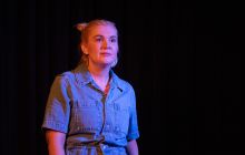 Writer/Performer Kirby Lunn is pictured in a dark background, looking up, wearing a denim jumpsuit