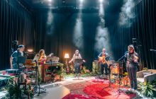 Five performers are light in a smoky atmosphere, with greenery, in a half circle